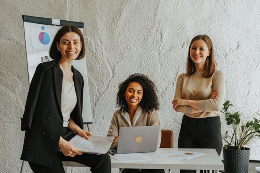 three women in professional clothing gathered around a table with a laptop on it and graphs in the background
